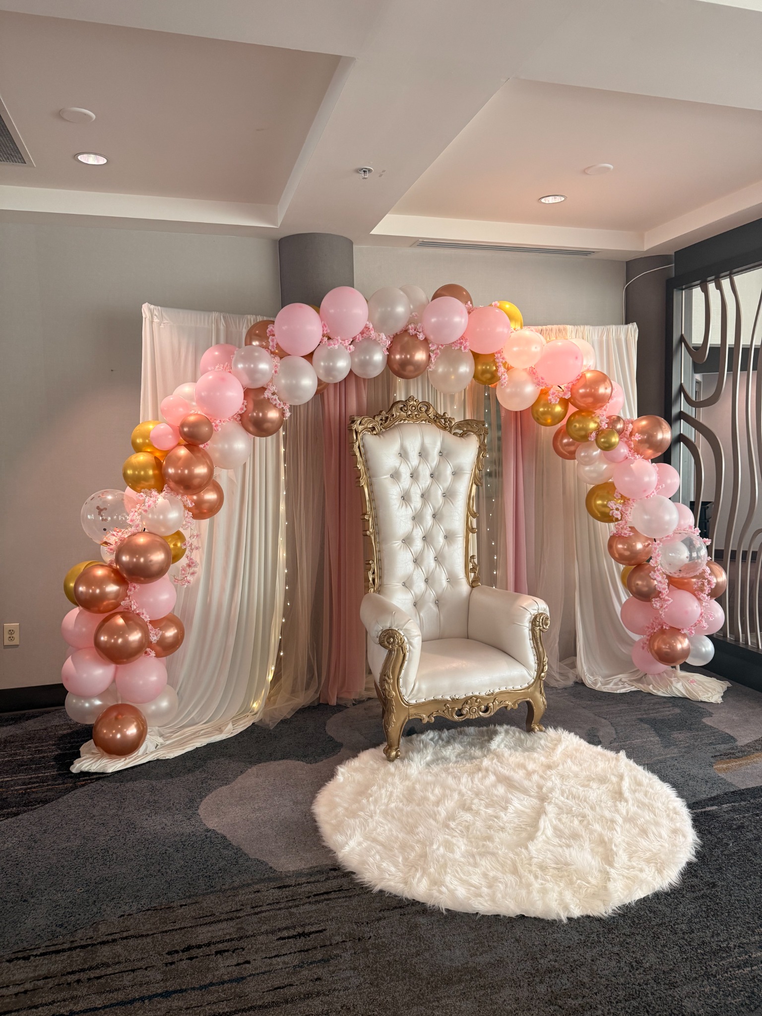 Pink, gold, and pearl balloon arch with draped backdrop and ornate throne chair